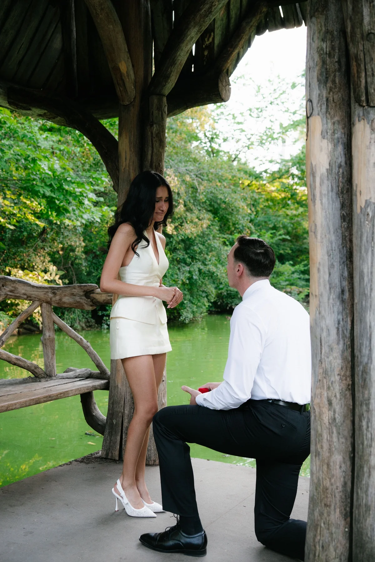 A man kneeling on one knee to propose to his partner beside a wooded pond in New York City